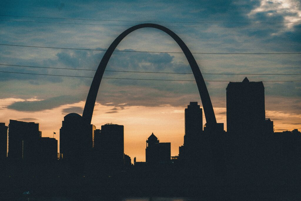 The St. Louis Gateway Arch is center in frame with the St. Louis skyline. There are clouds in the top half of the photo, giving way to a sunrise. 